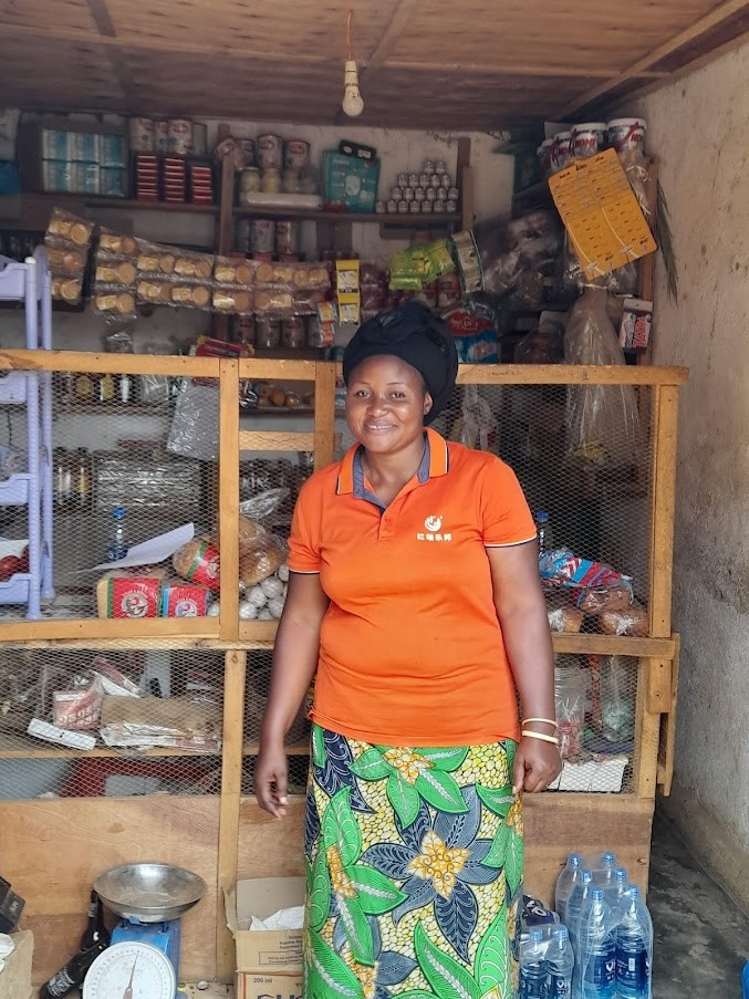 Ruth, stands smiling in her shop surrounded by produce, drinks and snacks
